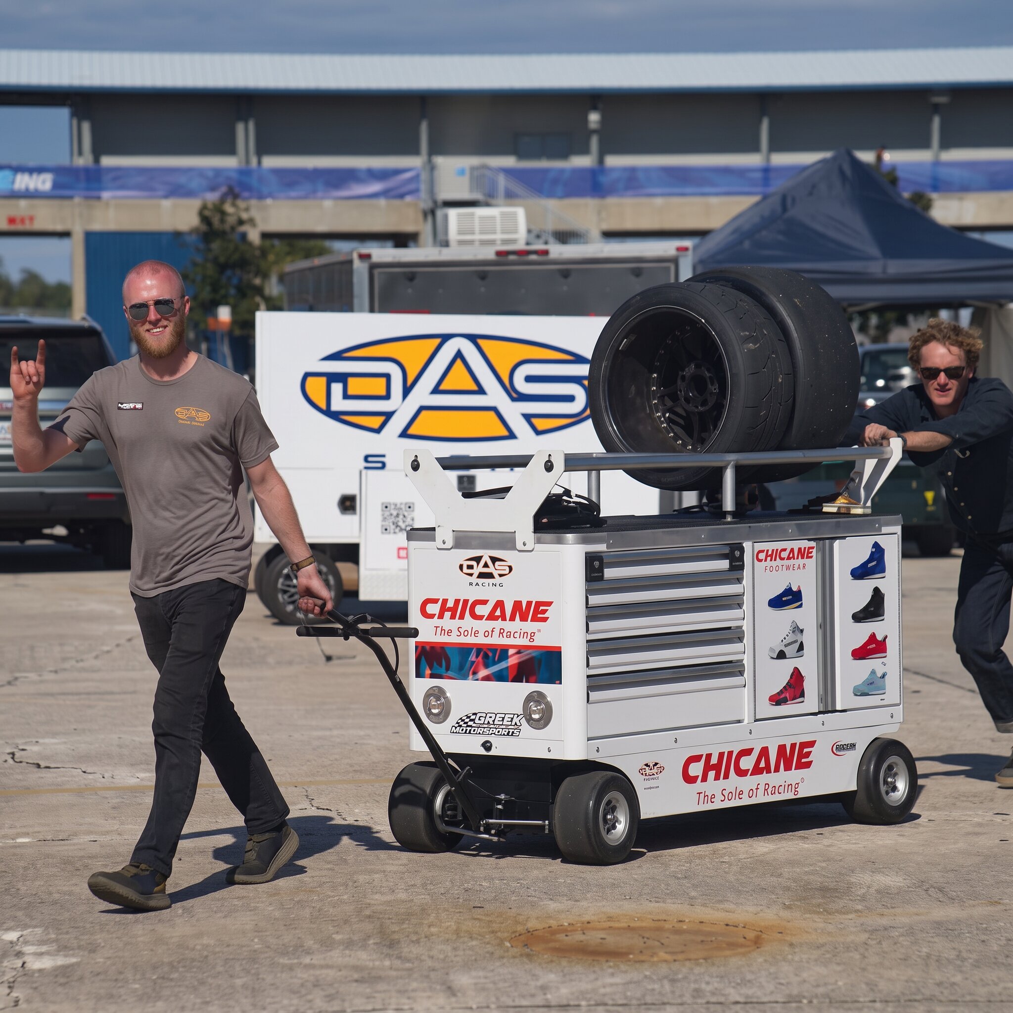 DAS Sport crew rolling a tool cart and slicks through the paddock in front of the team transporter
