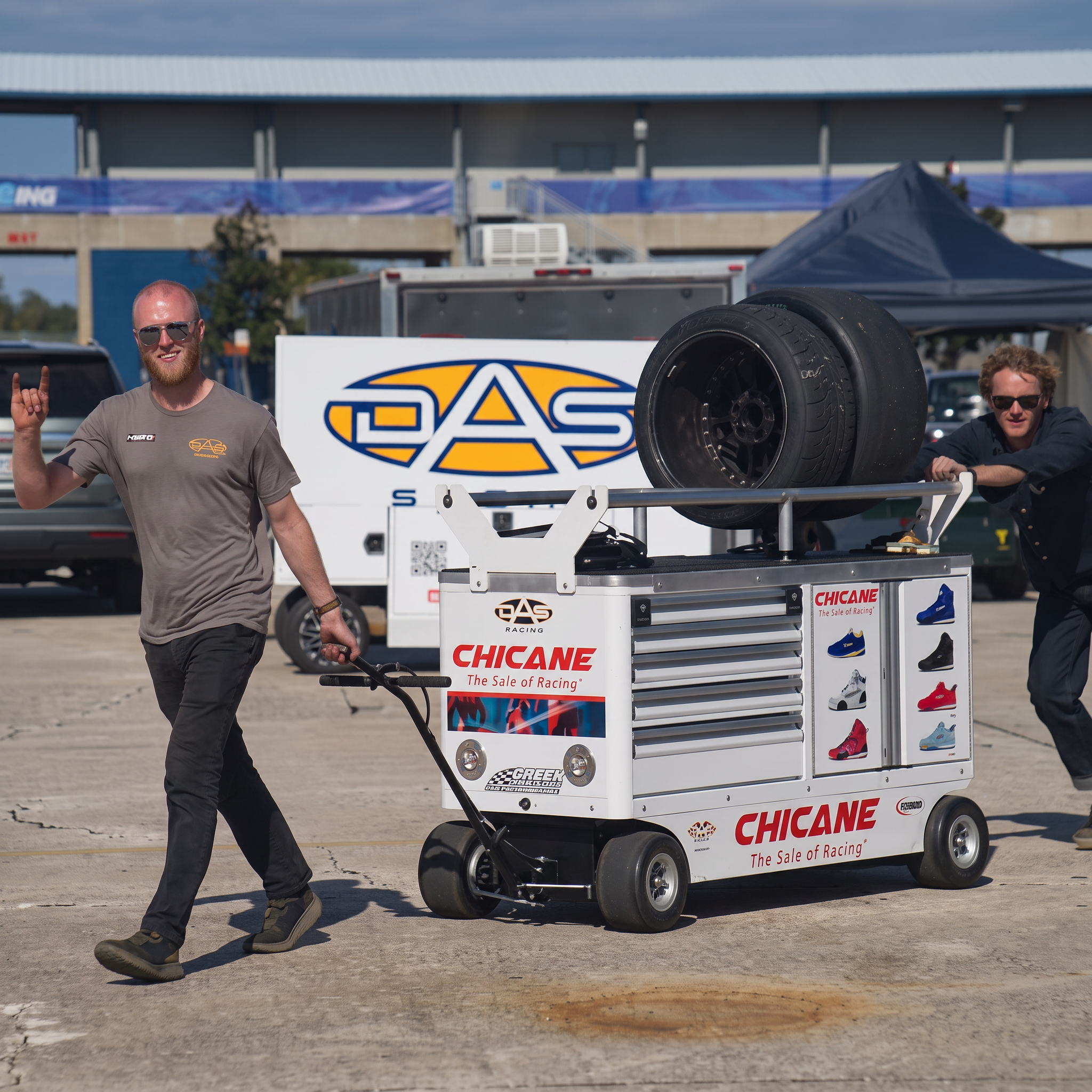 DAS Sport crew rolling a tool cart and slicks through the paddock in front of the team transporter